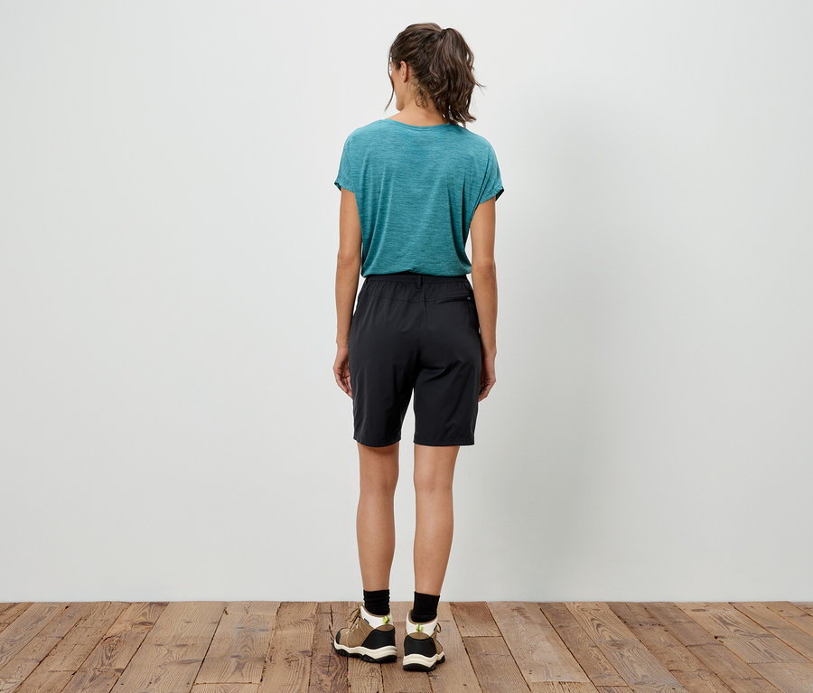Vue de dos d'une femme avec une queue de cheval portant un t-shirt turquoise, un short noir et des baskets sur un plancher en bois.