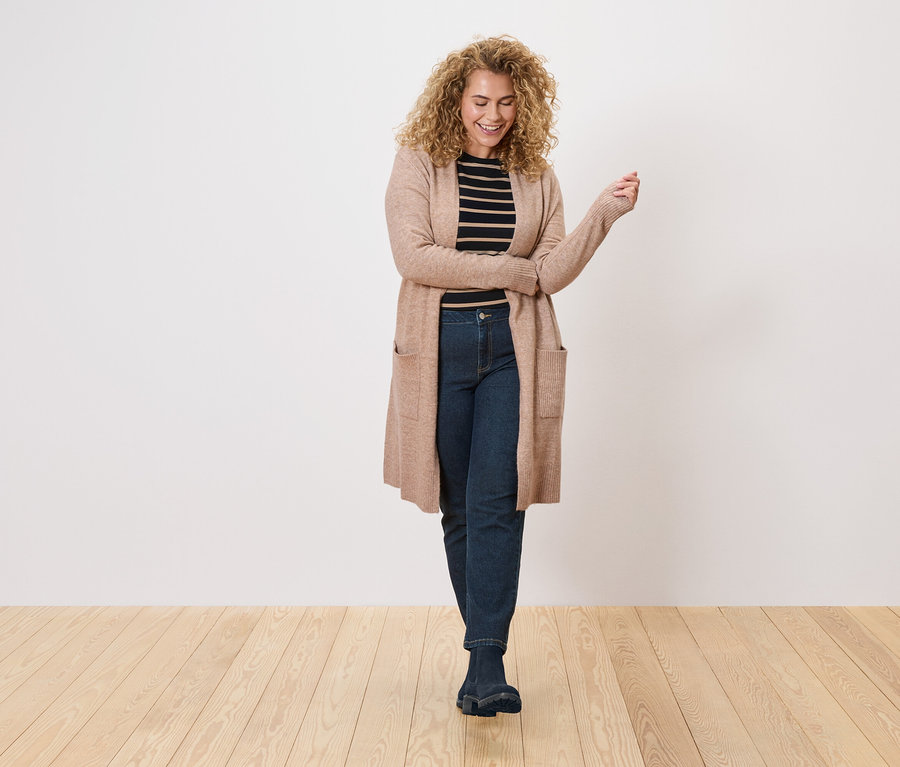Une femme aux cheveux bouclés porte un long cardigan marron clair sur un haut rayé noir et blanc et un jean foncé.
