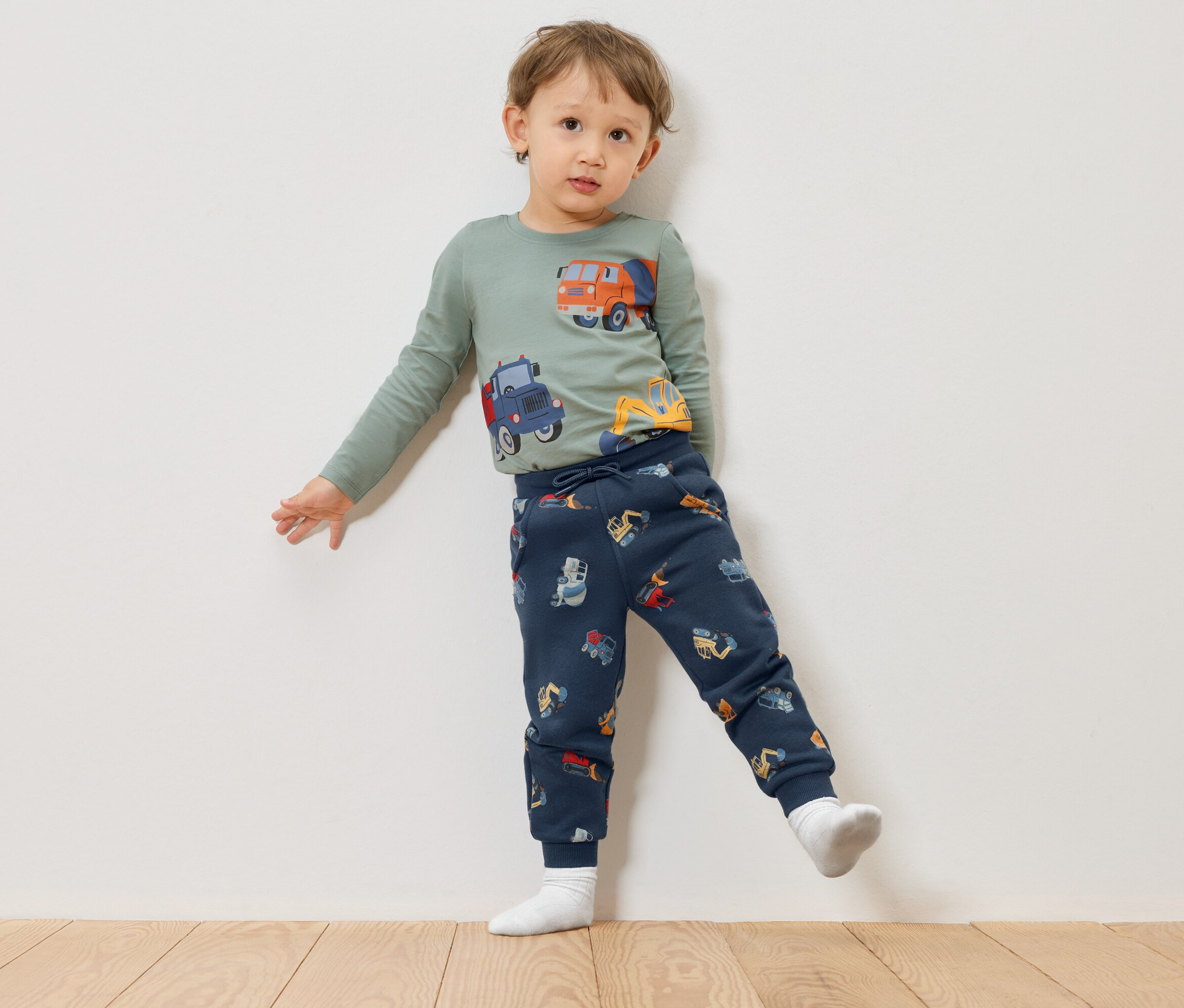Un enfant portant un t-shirt vert à manches longues avec des motifs de camions de chantier et un pantalon bleu marine avec des motifs de camions de chantier se tient devant un mur blanc.