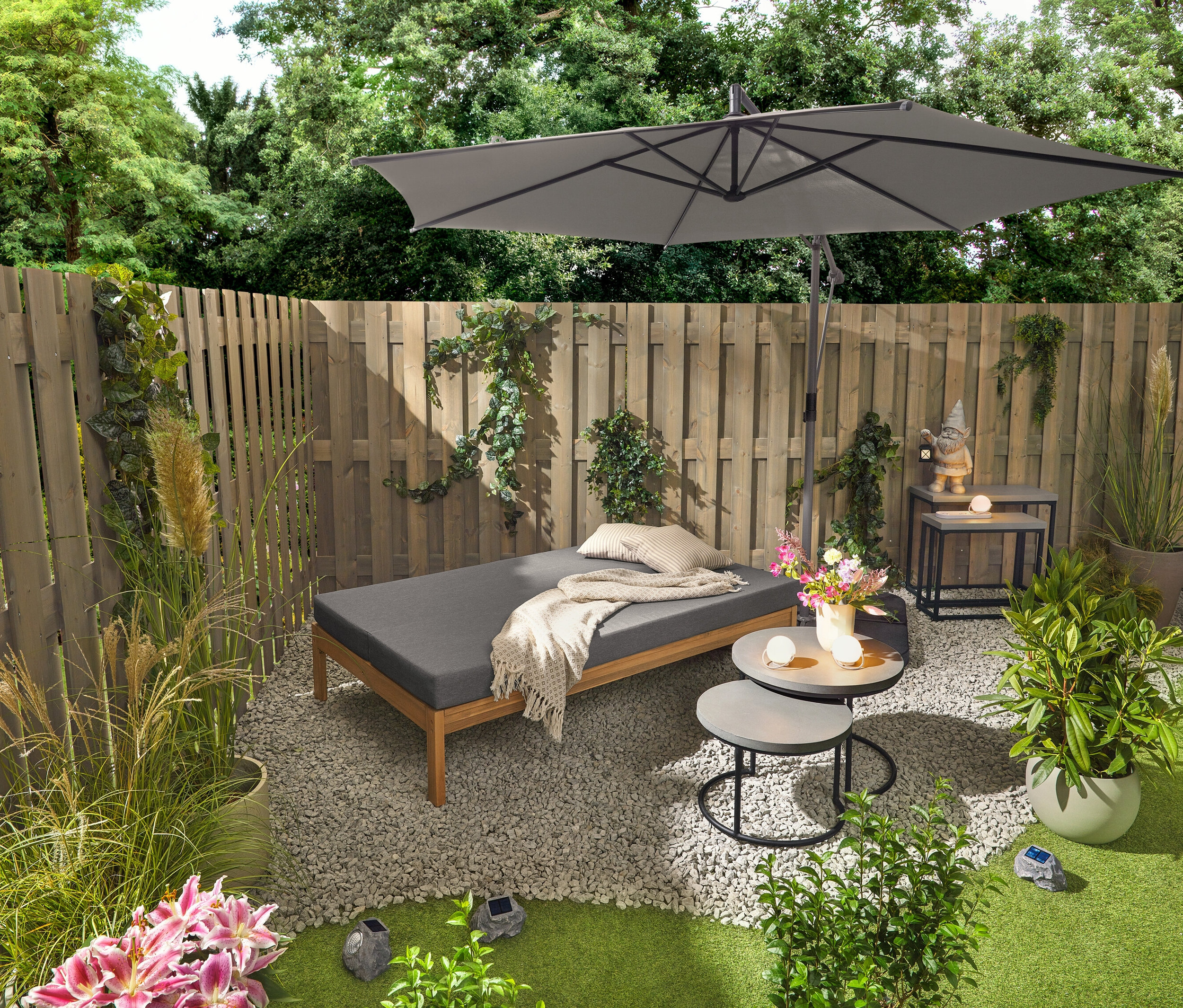 Un banc de jardin 2 en 1 servant de bain de soleil double est placé à côté de deux tables de jardin avec plateaux en polyciment (d'environ 50 cm et 60 cm de diamètre). Un nain de jardin avec lanterne LED se trouve sur l'une des 2 tables consoles de jardin avec plateaux en polyciment. Trois lampes de table à accu et trois pierres à LED solaires Premium sont également visibles.