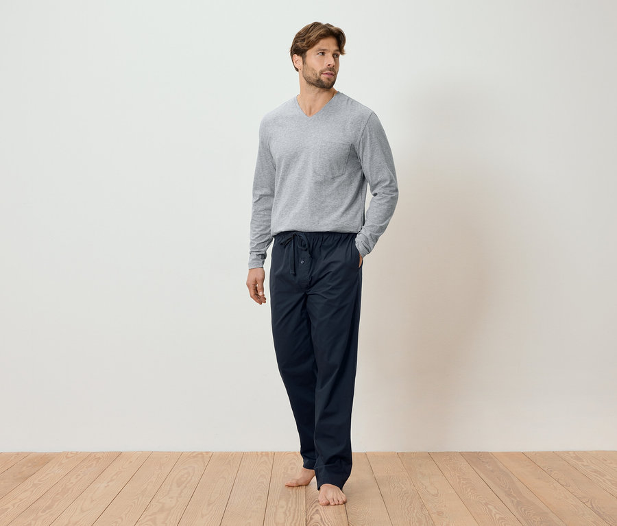 Un homme debout pieds nus sur un plancher en bois, portant un t-shirt à manches longues gris et un pantalon de pyjama bleu marine.