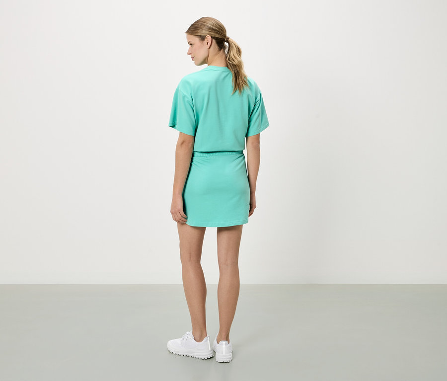 Une femme aux cheveux blonds, vue de dos, porte un haut à manches courtes vert menthe et une jupe assortie, ainsi que des baskets blanches.