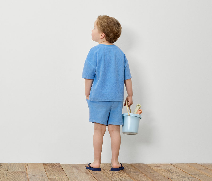 Garçon en ensemble éponge bleu tenant un seau bleu avec des jouets devant un mur blanc.