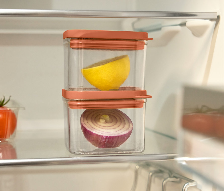 Dans le réfrigérateur, il y a deux récipients transparents avec des couvercles orange. Dans un récipient, il y a un demi-citron, dans l'autre un demi-oignon rouge. À gauche, il y a un bol de tomates.