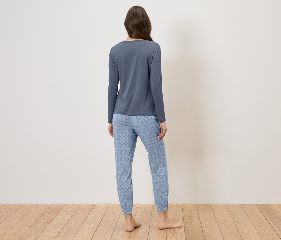 Une femme vue de dos portant un pyjama bleu à motifs, debout sur un plancher en bois devant un mur blanc.