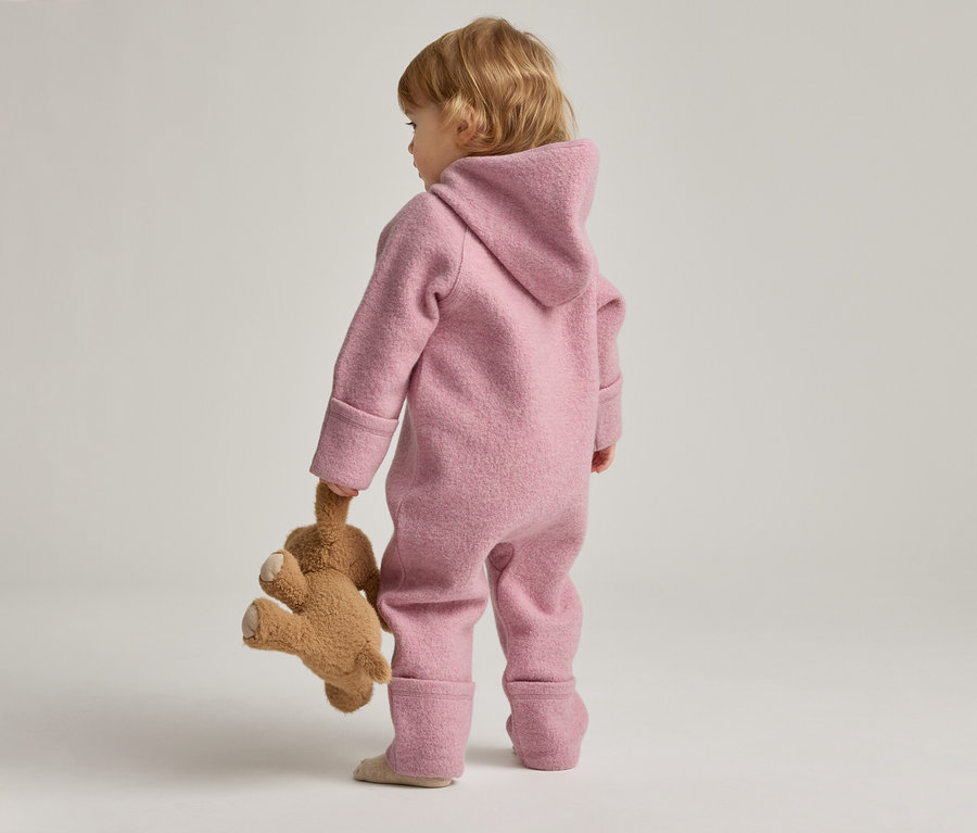 Vue de dos, un enfant en bas âge en combinaison rose à capuche tenant un ours en peluche.