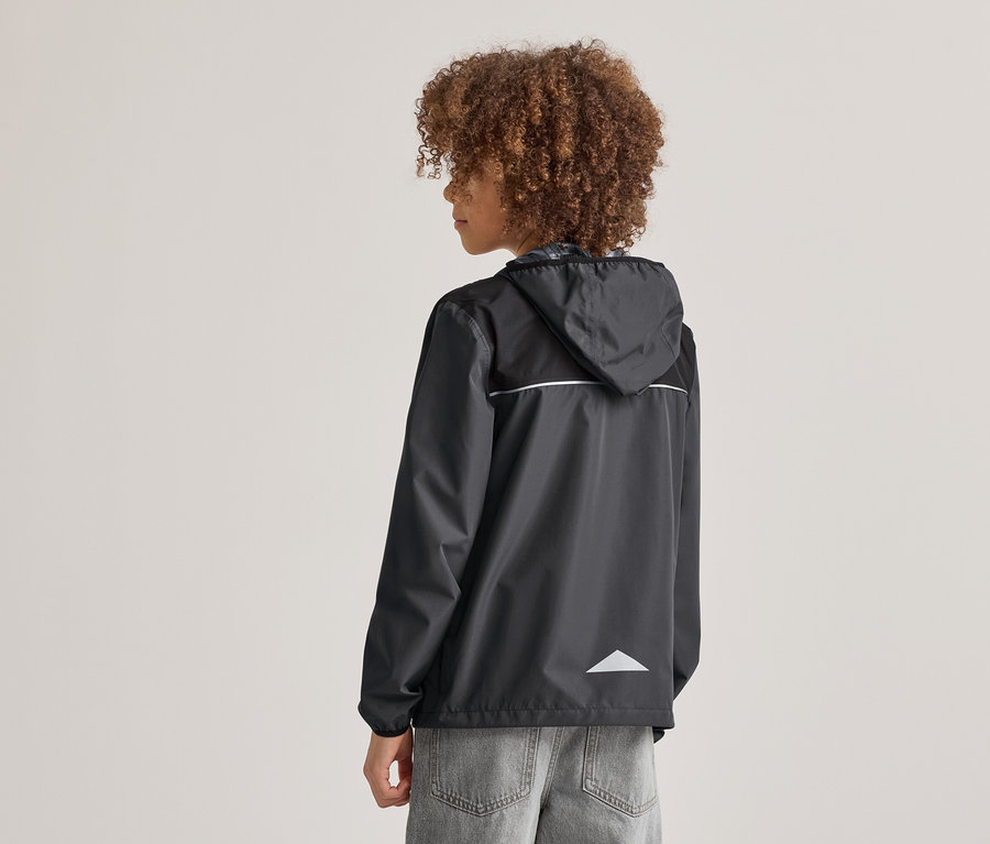 Personne aux cheveux bouclés vue de dos, portant un imperméable noir à capuche avec des détails réfléchissants et un jean gris.