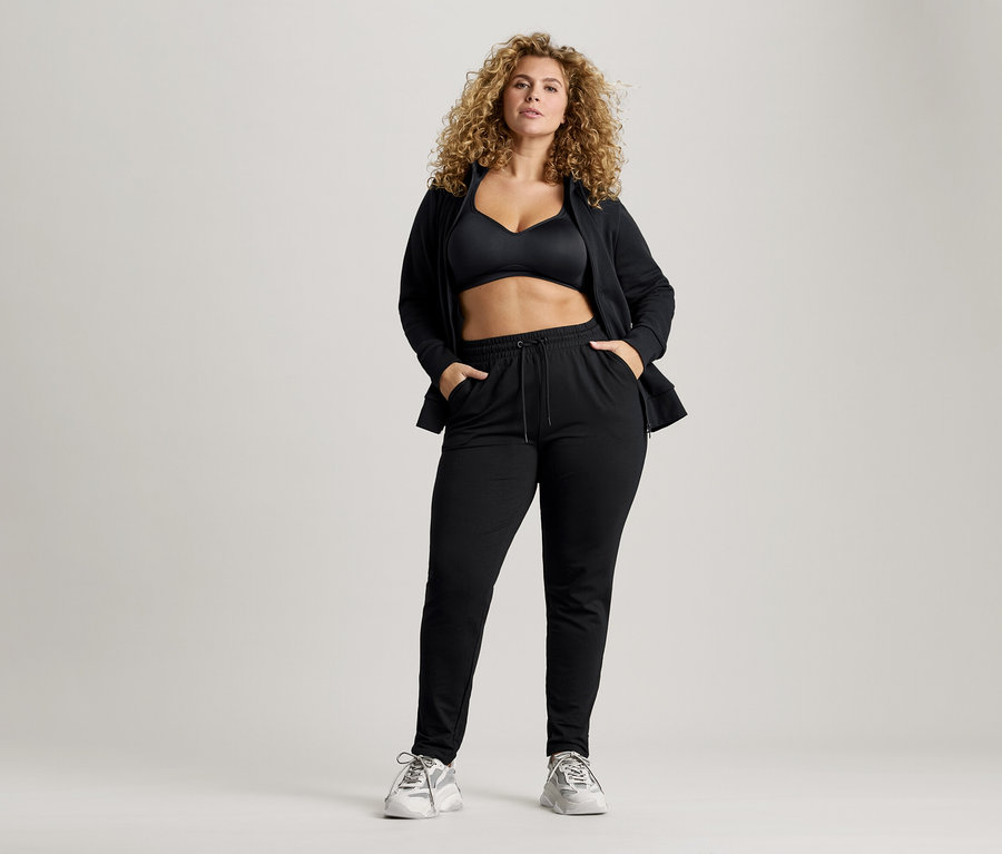 Femme aux cheveux bouclés portant un soutien-gorge de sport noir, une veste et un pantalon de survêtement avec les mains dans les poches.