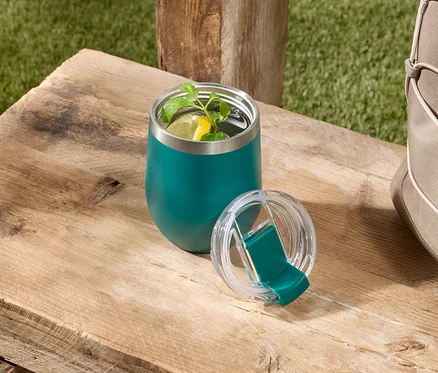 Un gobelet isotherme vert contenant une boisson avec du citron et de la menthe est posé avec son couvercle sur une table en bois à côté d'un sac beige.