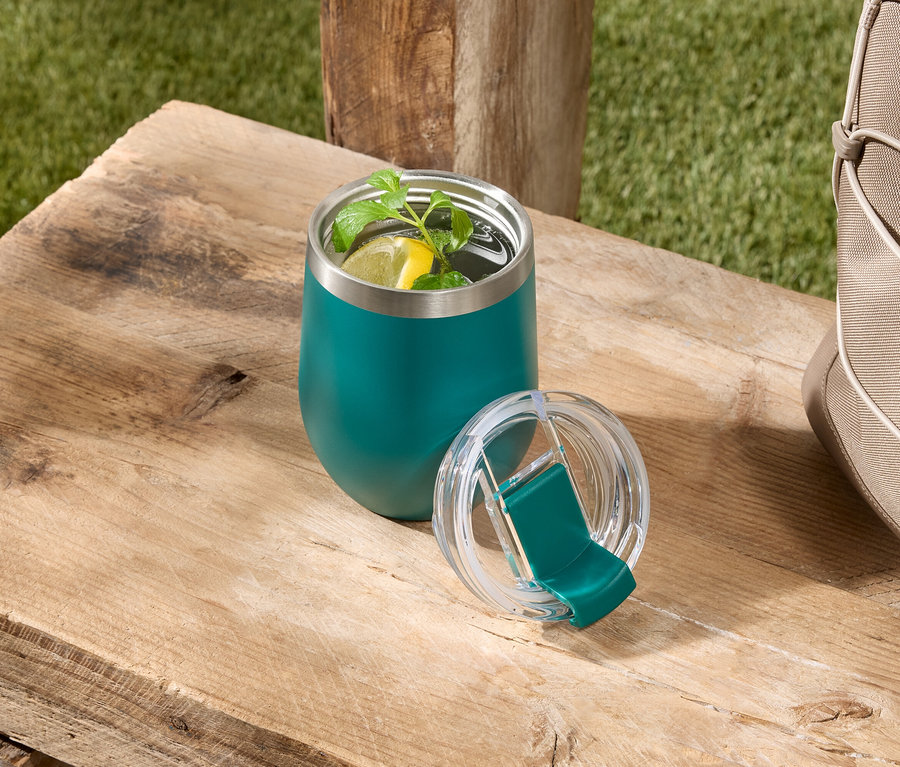 Un gobelet isotherme vert contenant une boisson avec du citron et de la menthe est posé avec son couvercle sur une table en bois à côté d'un sac beige.