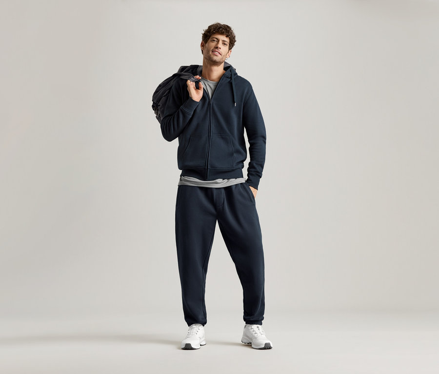 Homme portant un sweat à capuche bleu marine et un pantalon de jogging bleu marine, avec un sac sur l'épaule.