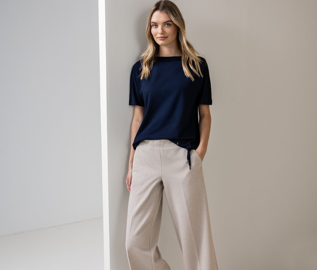 Une femme pose contre un mur, portant une chemise à manches courtes Street One bleu marine et un pantalon beige.