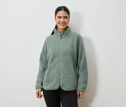 Une femme souriante avec une queue de cheval porte une veste en polaire verte zippée et un legging noir sur fond blanc.