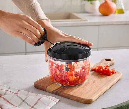 Des mains actionnent un hachoir manuel avec un récipient transparent rempli de légumes rouges hachés. Le hachoir est posé sur une planche à découper en bois avec d'autres légumes hachés. Un torchon à carreaux et une courgette sont sur le comptoir.