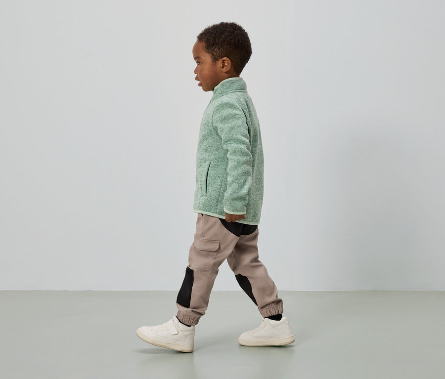 Un garçon à la peau foncée marche, portant une veste polaire verte, un pantalon cargo avec des empiècements noirs et des baskets crème.