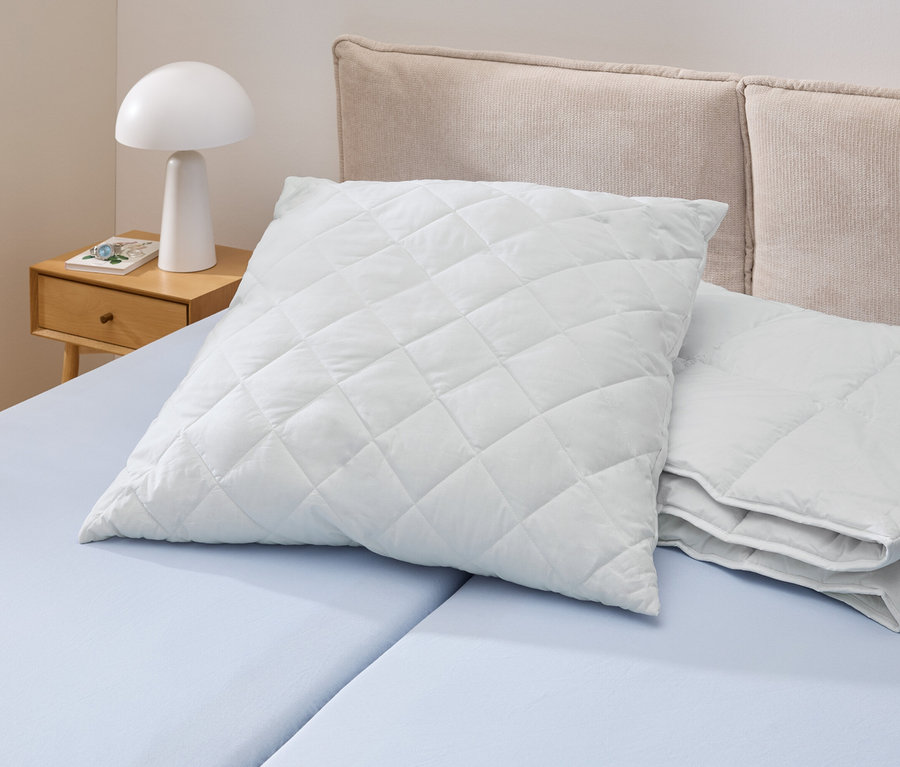 Un oreiller Cell Solution® blanc matelassé (90 x 70 cm) est posé sur un lit bleu clair. À côté se trouvent d'autres oreillers Cell Solution® (80 x 80 cm, 100 x 65 cm).