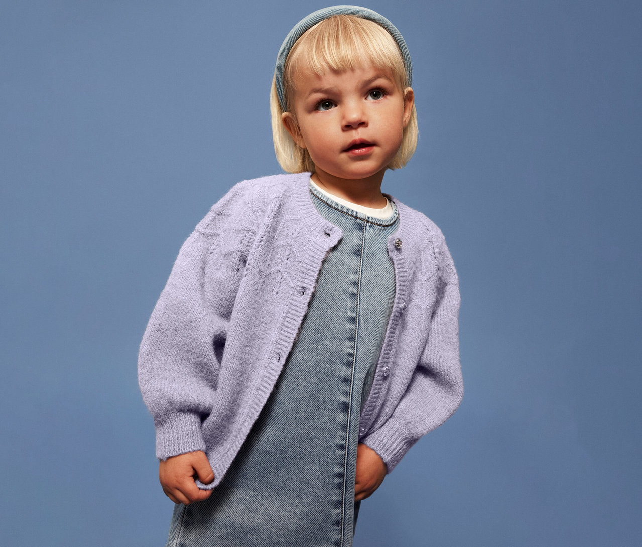 Une fille blonde porte un gilet en maille lilas clair NAME IT sur une robe en jean, devant un fond bleu.