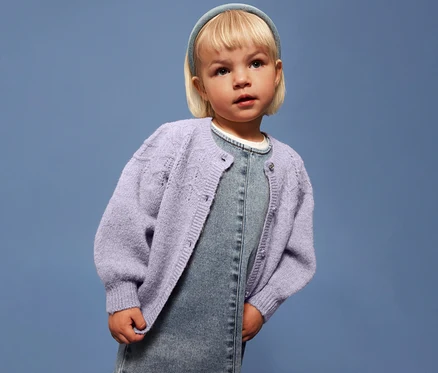 Une fille blonde porte un gilet en maille lilas clair NAME IT sur une robe en jean, devant un fond bleu.