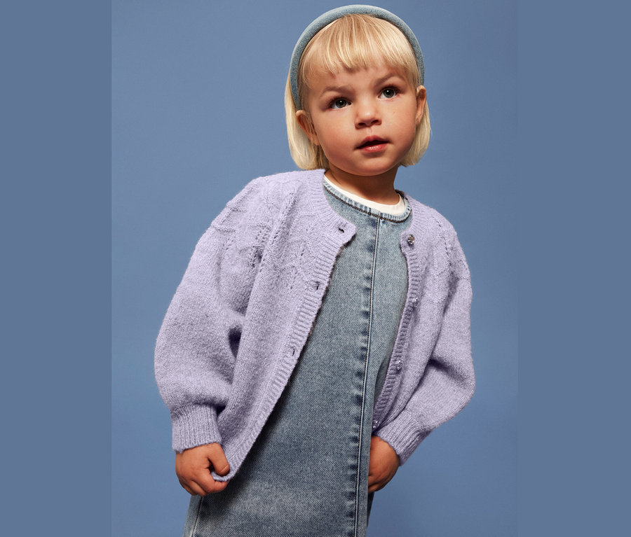 Petite fille blonde avec un bandeau bleu, un cardigan violet et une robe en jean sur un fond bleu.