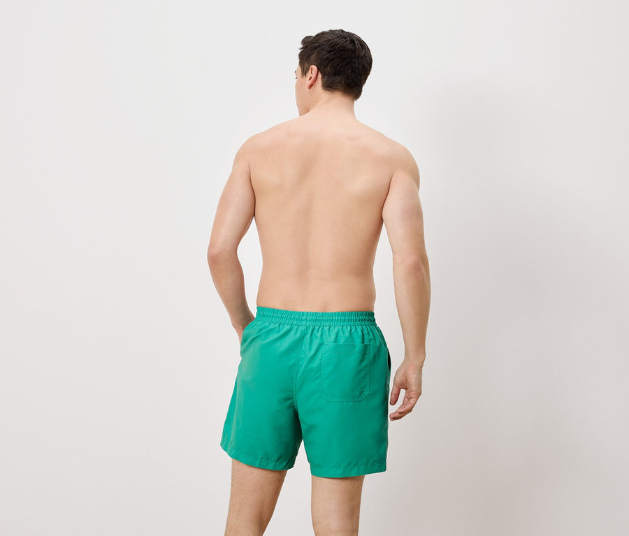 Un homme se tient dos à la caméra et porte un short de bain vert.