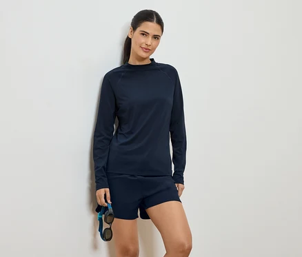 Une femme portant un haut à manches longues bleu marine et un short, appuyée contre un mur et tenant des lunettes de natation.