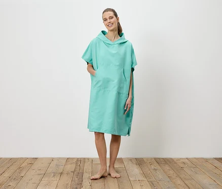 Une femme se tient pieds nus sur un plancher en bois et porte un poncho de bain turquoise à capuche avec une poche.