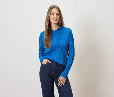 Une femme aux cheveux longs pose en pull bleu à capuche et jean foncé sur fond clair.