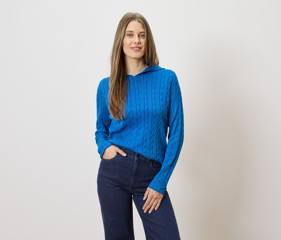 Une femme aux cheveux longs pose en pull bleu à capuche et jean foncé sur fond clair.