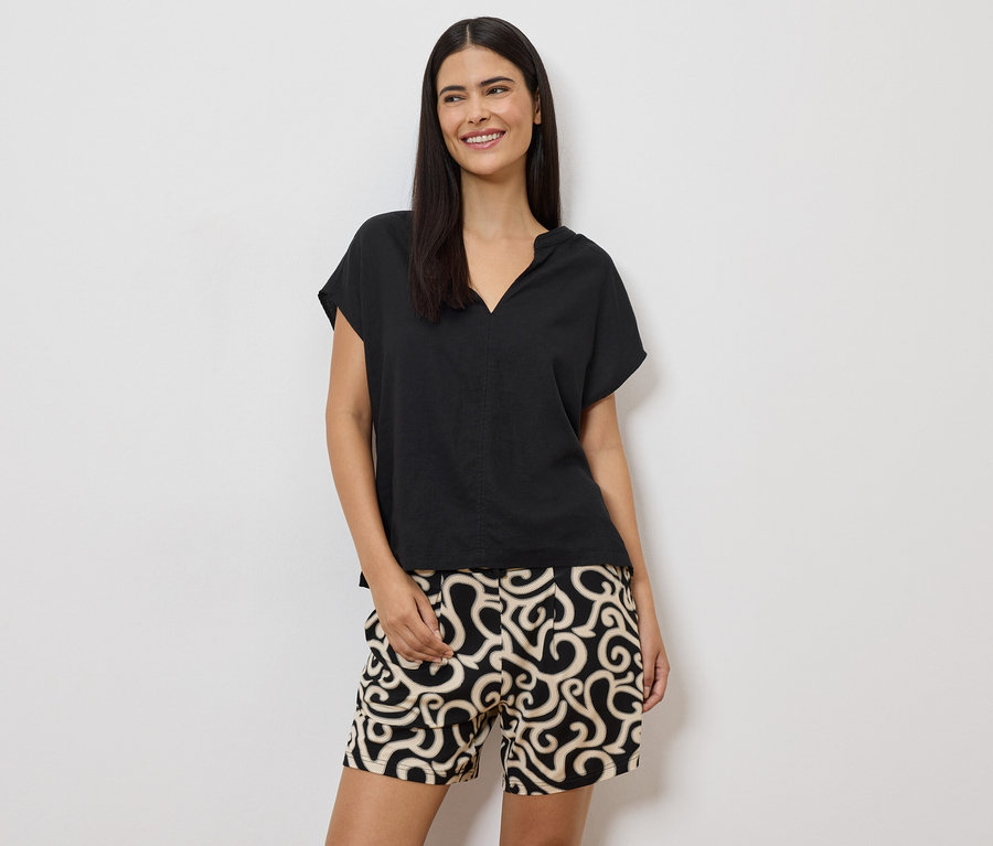 Une femme aux cheveux bruns mi-longs porte un chemisier noir et un short à motifs sur fond blanc.