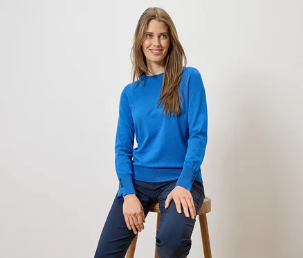 Une femme aux cheveux longs est assise sur un tabouret, portant un pull bleu et un pantalon bleu.