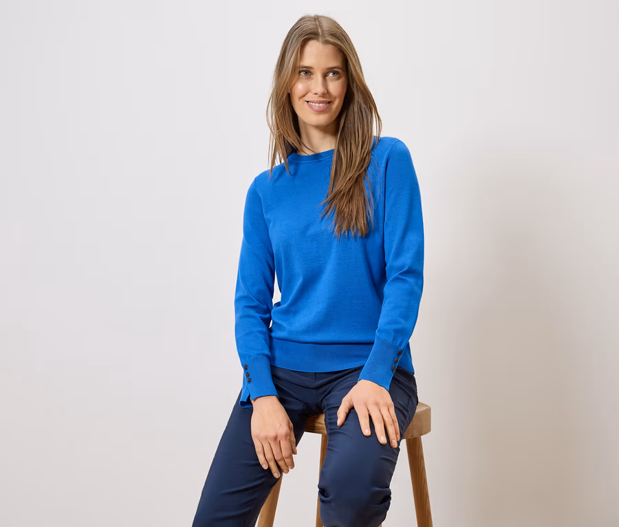 Une femme aux cheveux longs est assise sur un tabouret, portant un pull bleu et un pantalon bleu.