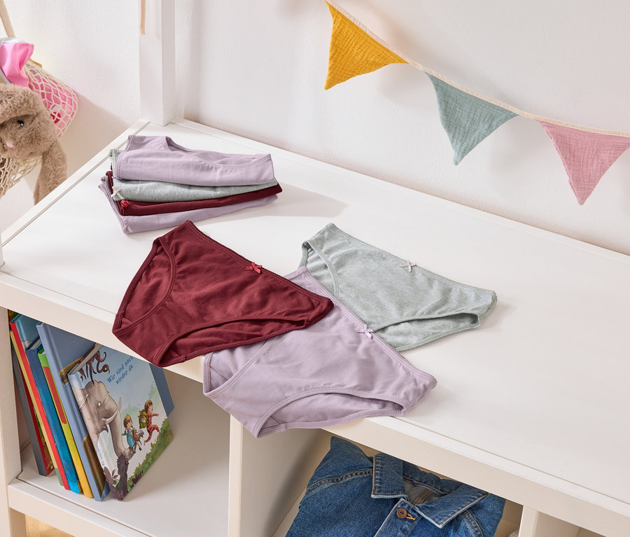 Plusieurs culottes de couleurs pastel sont posées sur une étagère blanche, avec des livres et une veste en jean en dessous. Une guirlande de fanions colorés est accrochée en arrière-plan.