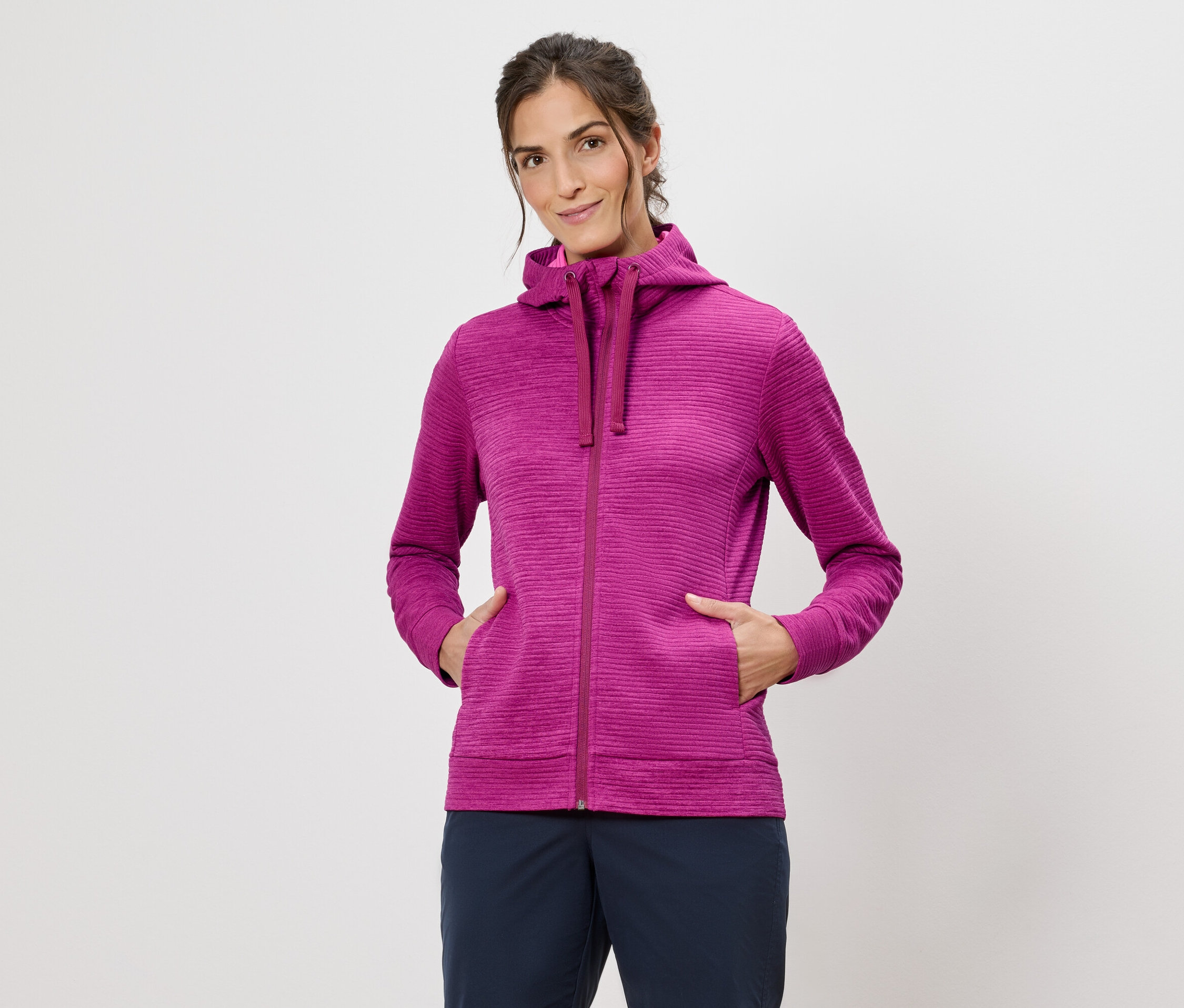 Femme portant un sweat à capuche rose, les mains dans les poches, sur un fond blanc.