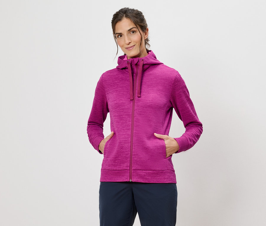 Femme portant un sweat à capuche rose, les mains dans les poches, sur un fond blanc.