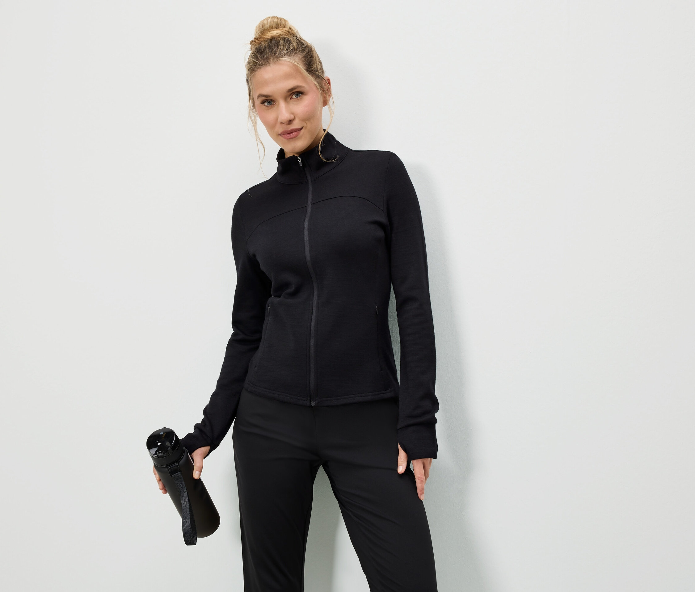 Femme en veste et pantalon noirs tenant une bouteille d'eau noire sur fond blanc.