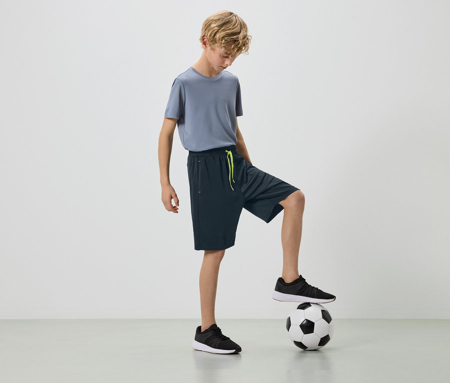 Un garçon blond en t-shirt gris et short foncé avec un pied sur un ballon de foot.