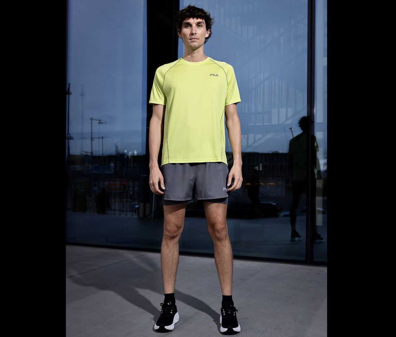 Homme en short gris et T-shirt technique jaune FILA devant une façade en verre. Il porte des chaussettes noires et des chaussures de course.