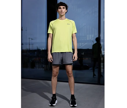 Un homme se tient devant une façade vitrée, portant un T-shirt technique pour homme FILA vert clair, un short gris et des chaussures de sport noires.