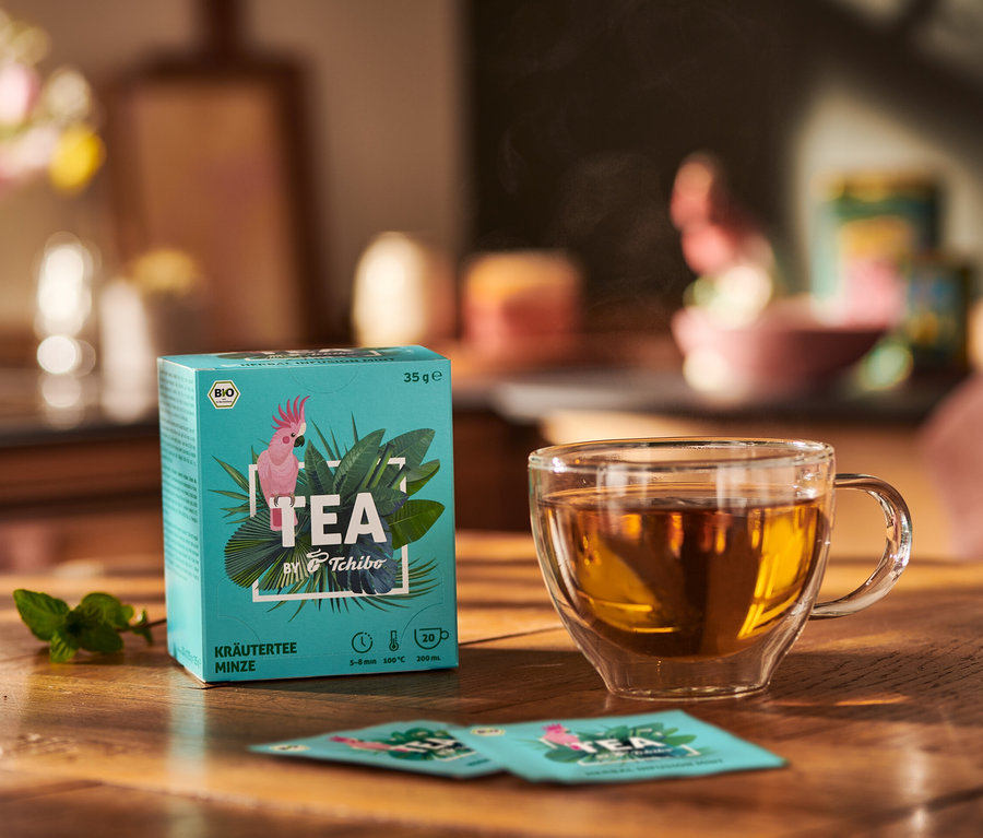 Paquet de BIO TEA by Tchibo Menthe à côté d'une tasse de thé et des feuilles de menthe sur une table en bois.
