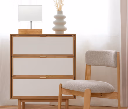 Une commode en bois clair avec trois tiroirs blancs et des poignées dorées, sur laquelle se trouvent une lampe de table blanche et un vase blanc avec des fleurs séchées. À côté se trouve une chaise en bois clair avec une assise rembourrée grise. La scène est sur un parquet en bois avec un tapis blanc, devant une fenêtre lumineuse avec des rideaux blancs.
