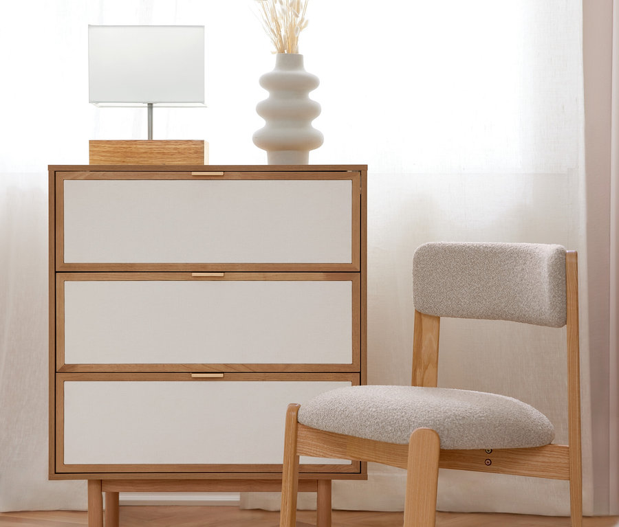Une commode en bois clair avec trois tiroirs blancs et des poignées dorées, sur laquelle se trouvent une lampe de table blanche et un vase blanc avec des fleurs séchées. À côté se trouve une chaise en bois clair avec une assise rembourrée grise. La scène est sur un parquet en bois avec un tapis blanc, devant une fenêtre lumineuse avec des rideaux blancs.