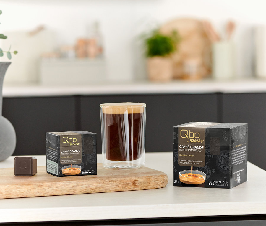 Sur une table, une tasse de café et des paquets de Qbo Caffè Grande ESPÍRITO SÃO PAULO de 8 et 27 capsules.