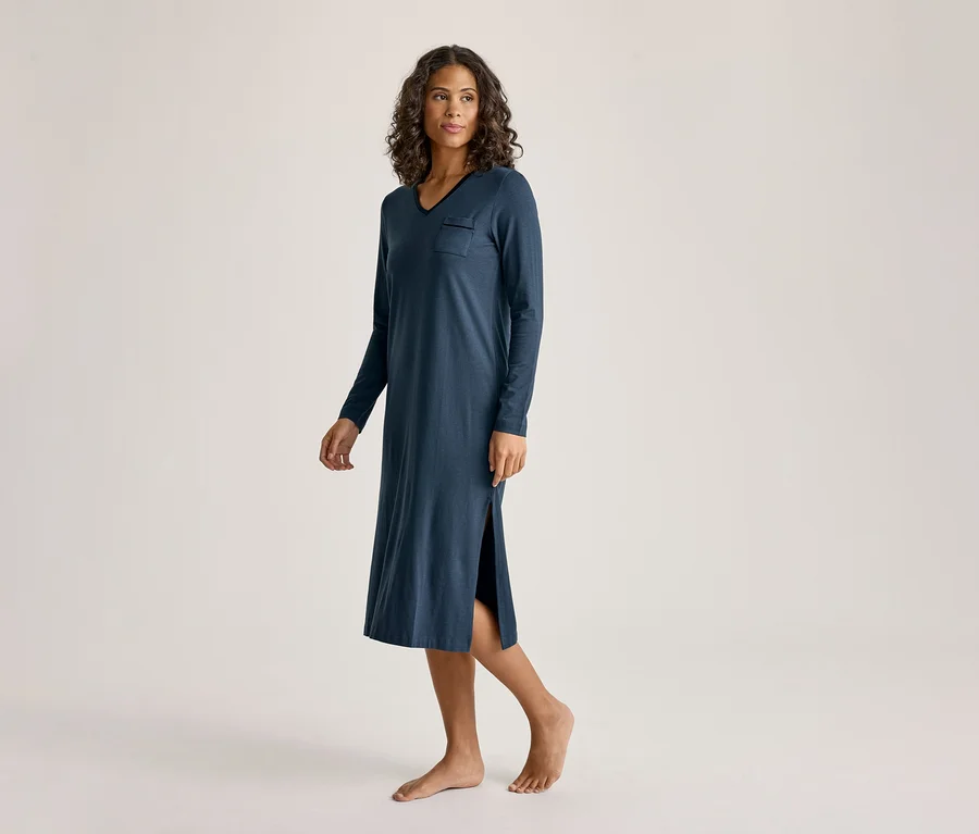Une femme aux cheveux bouclés porte une chemise de nuit bleu foncé à manches longues, col en V et poche poitrine, avec une fente latérale. Elle est pieds nus et regarde vers la droite.