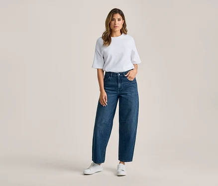 Femme debout, vêtue d'un t-shirt blanc, d'un jean large bleu foncé et de baskets blanches. Sa main droite est dans la poche de son jean.
