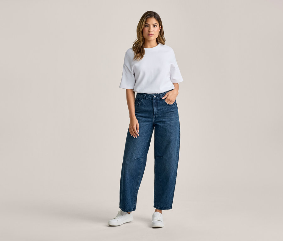 Femme debout, vêtue d'un t-shirt blanc, d'un jean large bleu foncé et de baskets blanches. Sa main droite est dans la poche de son jean.