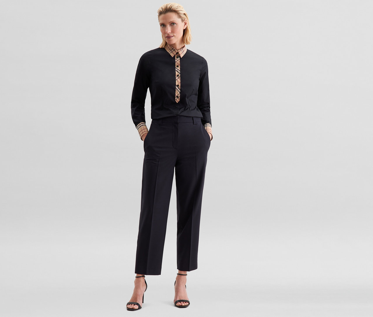 Femme debout, les mains dans les poches, portant un chemisier en popeline à carreaux Seidensticker noir, un pantalon noir raccourci et des sandales noires à talons hauts.