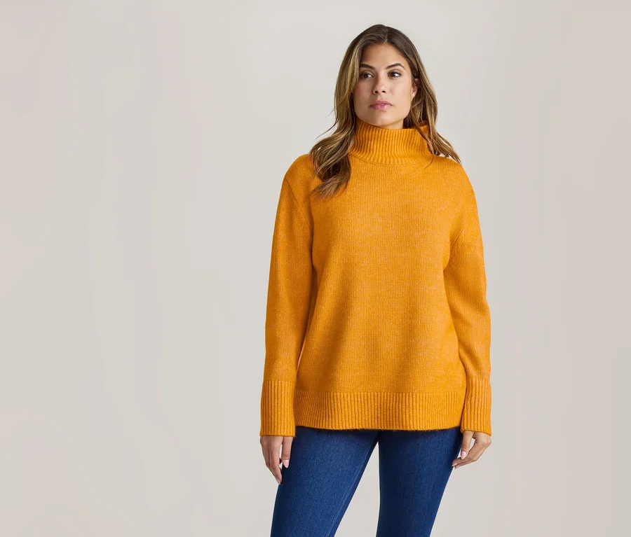 Une femme portant un pull à col roulé orange et un jean bleu se tient devant un fond clair, regardant légèrement vers la droite.