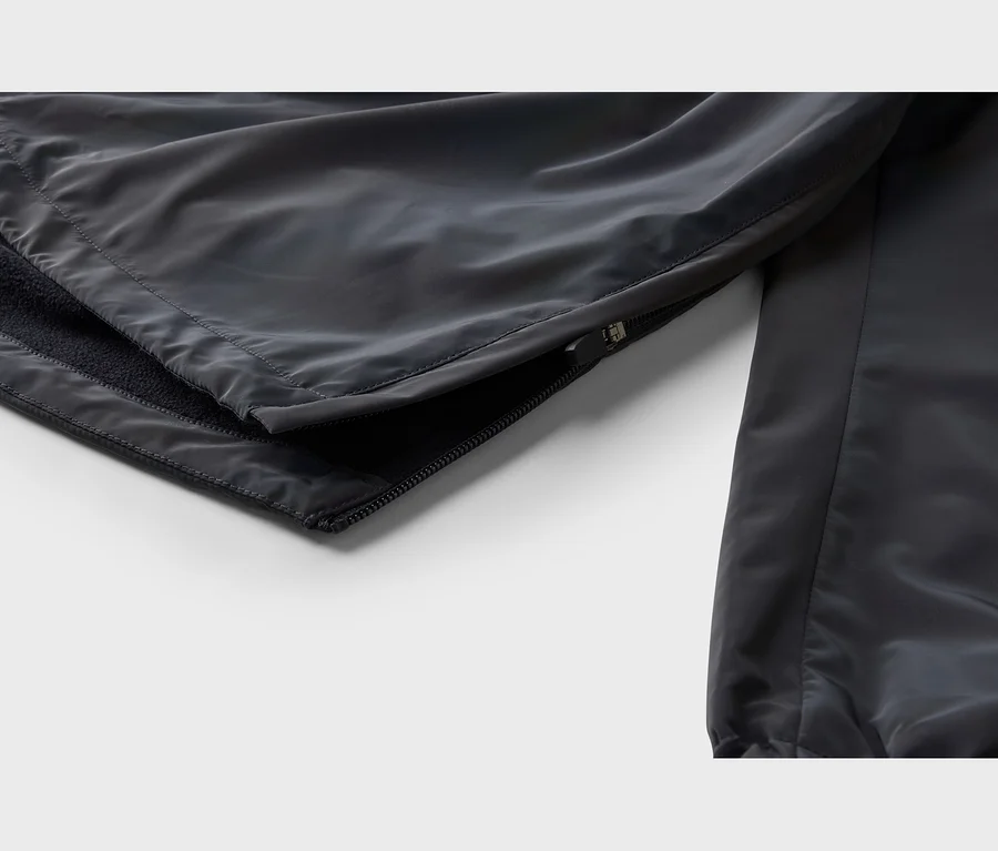 Gros plan d'un vêtement gris foncé, montrant une couture latérale partiellement ouverte révélant une doublure en polaire noire. Le tissu extérieur est lisse et légèrement brillant.