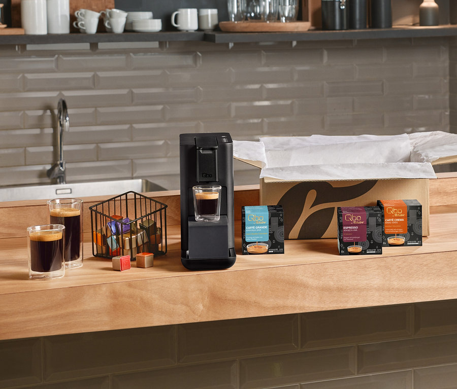 Machine à café noire Qbo Iconic, Dark Chrome sur un comptoir en bois, avec un verre de café en dessous. À côté, deux autres verres de café, un panier de capsules Qbo et des capsules en vrac. À droite, des paquets ouverts de capsules Qbo Caffè Grande KIRINYAGA BAYA, Qbo Espresso et Qbo Caffè Minas Verde.