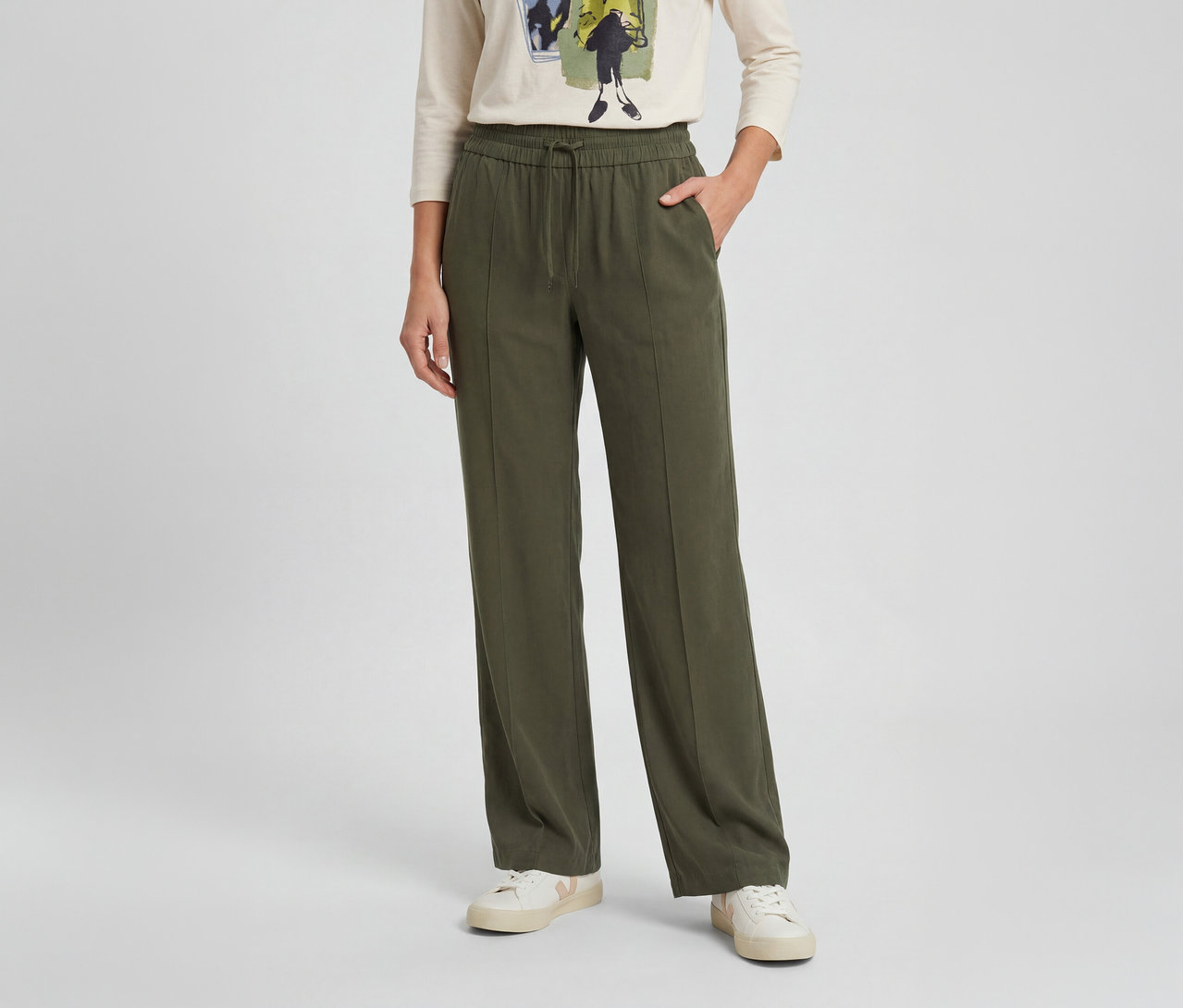 Personne portant un pantalon large vert olive avec cordon de serrage à la taille, des baskets blanches et un t-shirt clair à manches longues. Une main est dans la poche du pantalon.